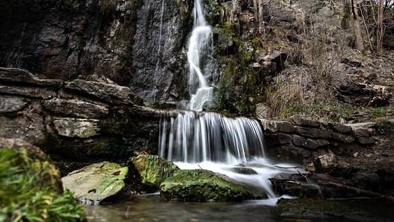 An einer Felswand fließt ein Wasserfall über einen Felsvorsprung, rechts davon kahle Bäume. 