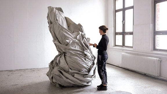 Ein Person steht vor einer einer Art zylindrischen Skulptur, die mit einem grauen Tuch umwickelt ist.