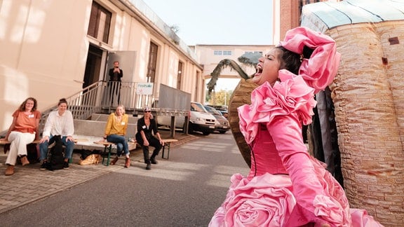 Eine junge Frau in einem knall-rosa Kostüm singt leidenschaftlich auf einem ehemaligen Fabrikgelände.