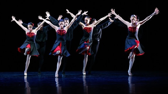 Frau in rot-schwarzen-Kleidern strecken synchron die Arme in die Luft und heben ein Bein.