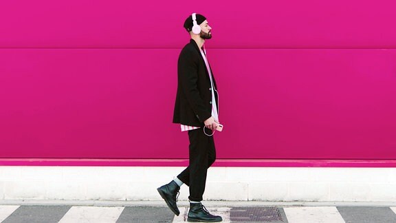 Junger Mann mit Kopfhörern und Smartphone geht an einer rosa Wand entlang.