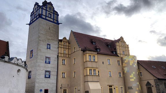 Ein historischer Bau mit Turm und Giebeln sowie Fensterumrandungen in blau und geelb erhebt sich auf einem Platz.