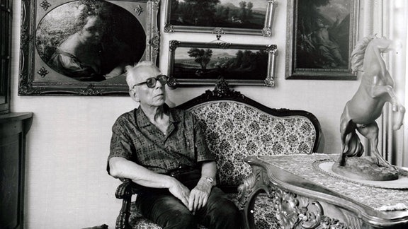 SchwarzWeiß-Bild: Der Sammler Friedrich Pappermann sitzt in seiner Wohnung auf einem Sofa vor einer Wand mit Gemälden.