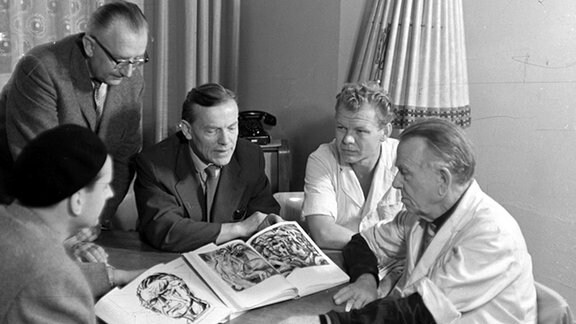Schwarz-Weiß-Foto: mehrere Männer stehen und sitzen um einen Tisch, auf dem aufgeschlagene Kunst-Kataloge liegen.