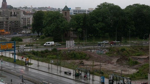 Blick auf eine Brachfläche mit Grabungen, im Hintergrund Stadt, davor eine Straßeund Haltestellen