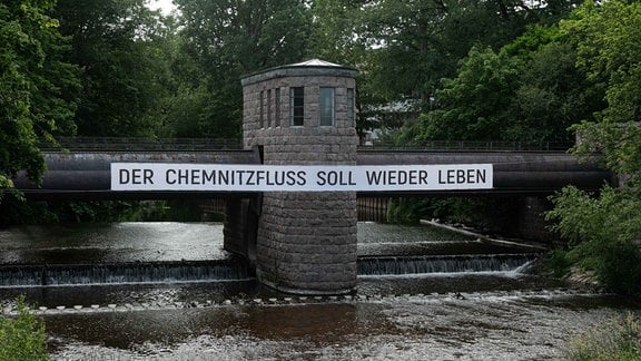 An einer Brücke über einen Fluss hängt ein Banner mit der Aufschrift: "Der Chemnitzfluss soll wieder leben"