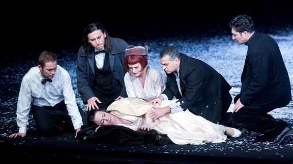 Auf einer Bühne liegt eine Frau auf dem Boden, um sie knien fünf Personen.