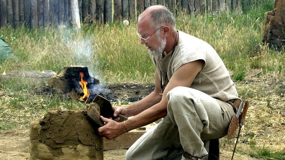 Ein Mann mit Brille und Backenbart kniet im Freien am Boden vor einem Feuer und hantiert mit Spatel und Lehm, um einen zylindrischen Ofen zu bauen.