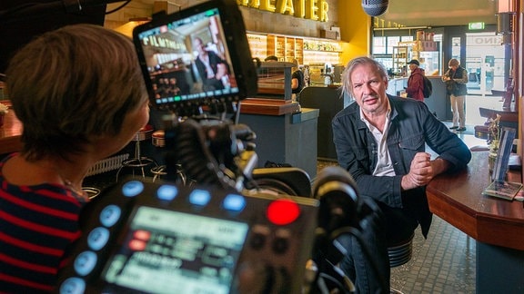 Regisseur Leander Haussmann im Interview mit dem MDR: Blick über eine Kamera hinweg auf einen Interviewpartner, der auf einem Hocker an einem Tresen sitzt, an der Wand leuchtet der Schriftzug Filmtheater