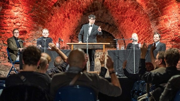 Veranstaltung beim Festival "Leipzig denkt" in der Moritzbastei, auf einer Bühne sitzen vier Personen an Tischen, in der Mitte steht ein Mann mit Anzug.