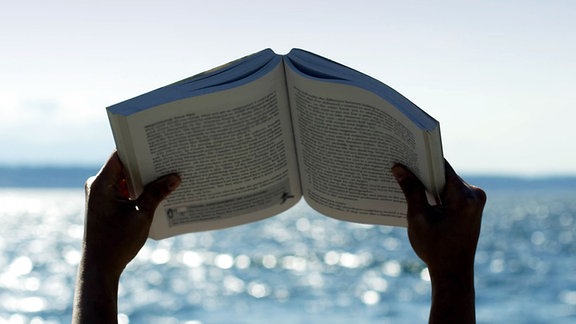 Eine Person hält ein aufgeschlagenes Buch gen Himmel. Im Hintergrund sieht man das Meer.