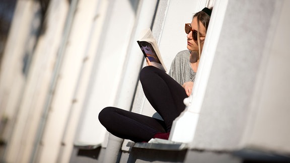 Eine Frau sitzt auf der Fensterbank im Sonnenschein und liest ein Buch