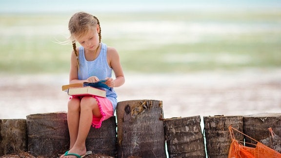 Ein Kind sitzt auf einer Buhne am Strand und hat ein Buch auf dem Schoß