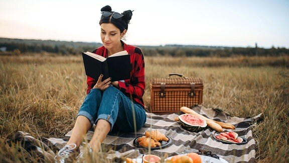 Eine Frau liest in einem Buch während eines Picknicks auf einer Wiese.