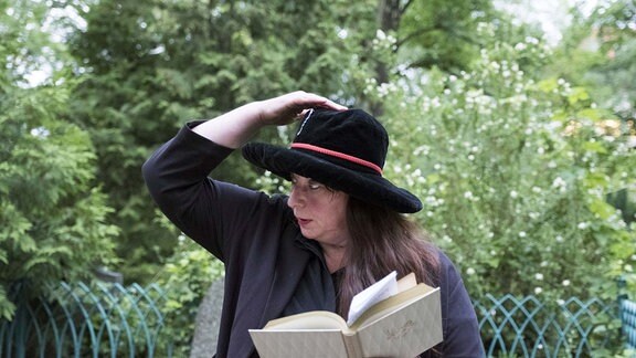 Eine Frau mit langen Haaren  hält in der einen Hand ein Buch, mit der andere fasst sie an den schwarzen Hut auf ihrem Kopf.  