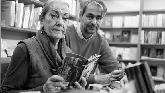 Eine Frau und ein Mann sitzen in einer Buchhandlung und schauen in die Kamera.