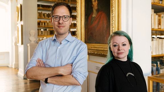 Ein Mann mit Brille, kurzen Haaren und hellblauem Hemd und eine Frau mit schwarzer Kleidung und türkisen Haaren stehen in einer historischen Bibliothek und blicken in die Kamera.