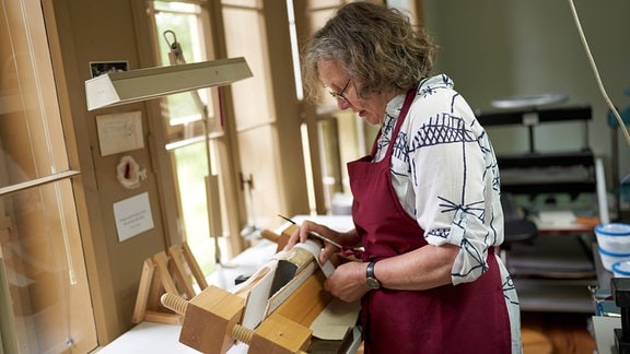 Eine Frau mit roter Schürze, grauem Haar und Brille beugt sich mit Werkzeug in den Händen über einen historischen Buchrücken.