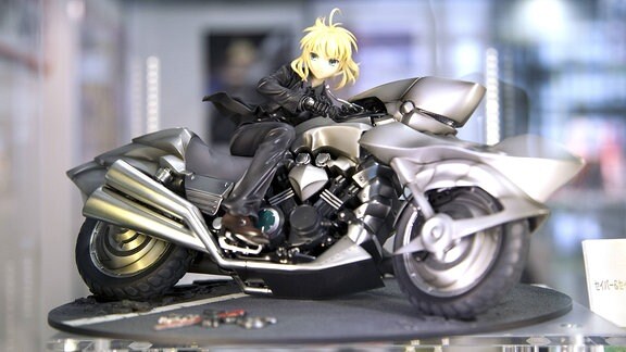 Ein silberfarbenes Spielzeugmotorrad mit einer Anime-Figur in einem schwarzen Anzug darauf steht in einer Vitrine 