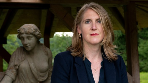 Juliane Stückrad - Blonde Frau mit halblangen Haaren und dunklem Jackett steht vor Statue