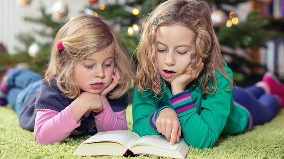 Zwei blonde Mädchen lesen ein Buch vor einem Weihnachtsbaum.