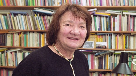 Eine Frau mit schwarzem Pullover und dunklem Haar steht vor einer Bücherwand und lächelt in die Kamera.