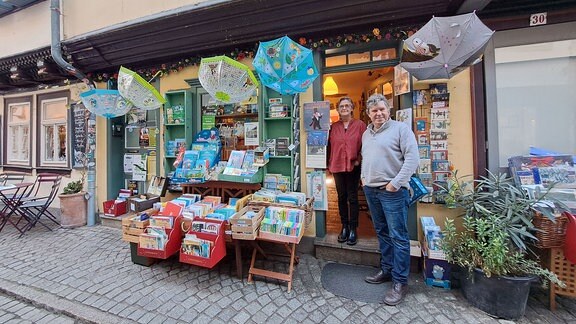 Eine Frau und ein Mann stehen vor einer Buchhandlung in einem Fachwerkhaus. Davor stehen Boxen mit bunten Büchern.
