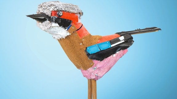 Skulptur eines Vogels mit langen Beinen und gelben Stiefelchen aus verschiedenen Materialien.