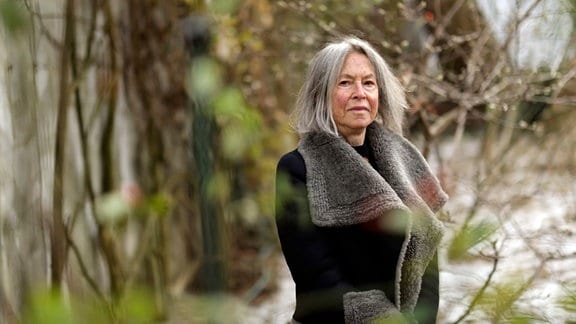 Die US-amerikanische Lyrikerin und Literaturnobelpreisträgerin Louise Glück steht hinter Zweigen eines Busches. 