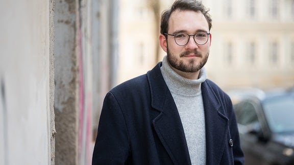 Der Autor Lukas Rietzschel: Ein Mann mit kurzen dunklen Haaren, dunkler Brille und Bart steht vor einer Häuserzeile.