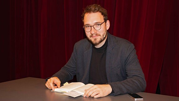 Lukas Rietzschel, ein junger Mann mit Brille sitz vor einem Buch an einem Tisch