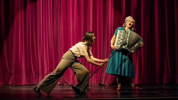 eine Frau spielt Akkordeon und singt dazu auf einer Bühne, eine andere Frau hält ihr ein Mikrofon entgegen