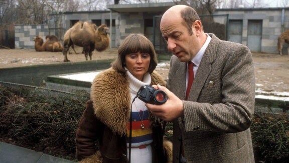Manfred und Ottilie Krug besuchen zusammen den Berliner Zoo. Sie schauen gemeinsam auf eine Kamera, hinten sieht man ein Kamel stehen.