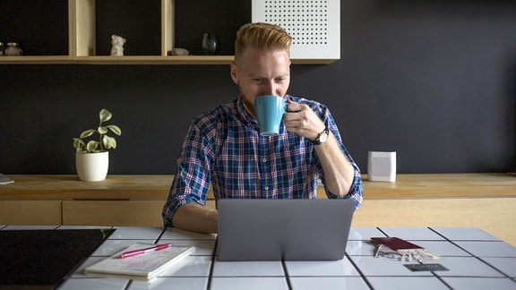 Mann arbeitet zu Hause an einem Laptop und trinkt dabei einen Kaffee.