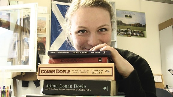 Eine Frau schaut lächelnd über einen Stapel mit Bücher über Sherlock Holmes und Arthur Conan Doyle
