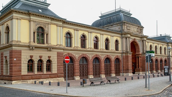 Flacher Bau mit gelber Fassade, großem Torbogen in der Mitte und je fünf Toren und Fenstern zu jeder Seite des Eingangs