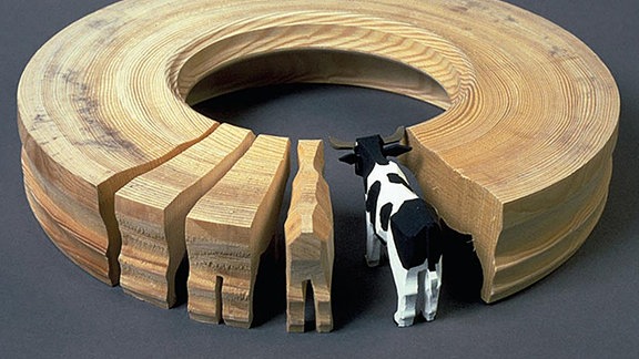 Ein Holzring, aus dem verschiedene Stück geschnitten wurden, an denen verschiedene Fertigungsschritte zu einer Kuh gezeigt werden.
