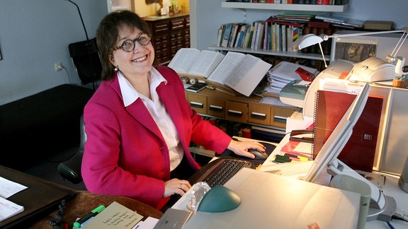 Eine Frau in pinken Jackett sitzt an einem Schreibtisch.