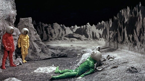 Szene aus dem Film ''Moon Zero Two''. Zwei Astronauten finden auf der Mondoberfläche einen am Boden liegenden Astronauten.