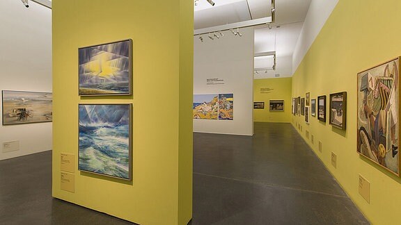 Blick in einen Ausstellungsraum mit gelben und weißen Wänden, an denen Gemälde hängen.