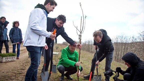 Eine Gruppe Männer pflanzt einen Baum, dabei hält einer den Baum, der andere gräbt mit einer Schauzfel ein Lock, ein anderer fotografiert.