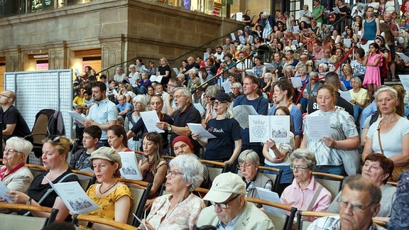 Blick in eine Menschenmenge auf einer Treppe, die Notenblätter in den Händen hält und singt