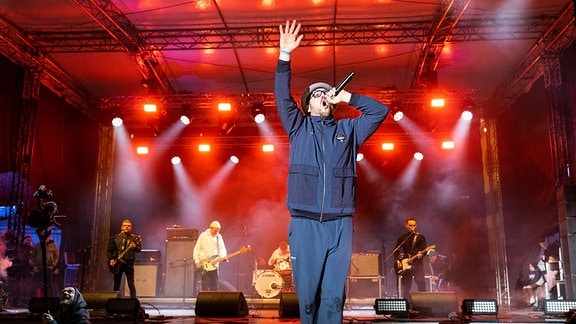 Eine Konzertbühne mit roten Scheinwerfern und einer Liveband, im Vordergrund ein Sänger mit Mikrofon, und Hut, der einen Arm in die Luft hebt