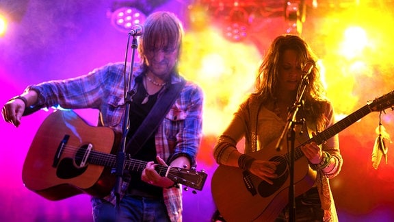 Eine Frau und ein Mann stehen mit Akustik-Gitarren an Mikrophonen auf einer Bühne.