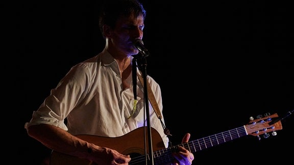 Ein Mann mit kurzen schwarzen Haaren und weißen Hemd steht mit einer Gitarre an einem Mirkofon und singt.