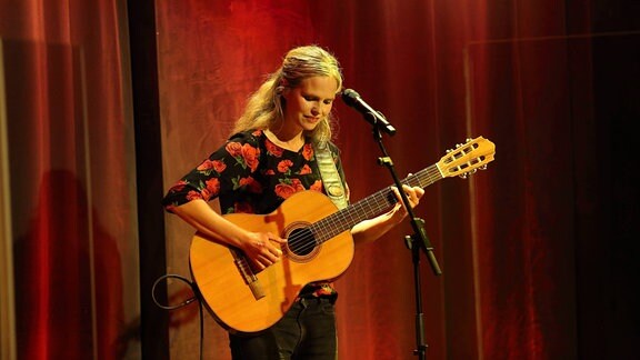Die Musikerin Dota Kehr steht mit ihrer Gitarre auf einer Bühne, im Hintergrund ein roter Vorhang.