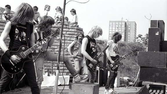 Vier Heavy-Metal Musiker auf der Bühne, hinter ihnen Kinder auf einer Mauer und Plattenbauten