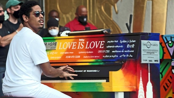 Matt Whitaker sitzt in weißen Sachen vor einem regenbogenbunten Klavier, auf dem er spielt, er trägt eine Sonnenbrille. Auf dem Klavier steht Love is Love