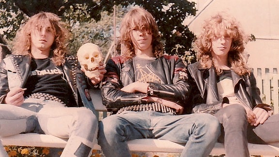 Drei Mitglieder der DDR-Metal-Band M.A.D. sitzen in Lederjacken auf einer Bank, einer hält einen Totenkopf in der Hand