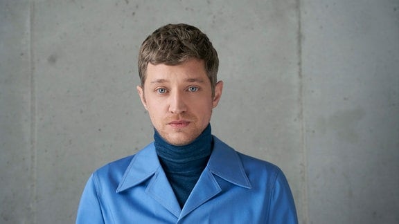 Portät des Musiker Maeckes in blauem Rollkragenpullover und hellblauem Jackett vor einer Betonwand.